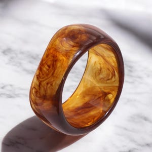 May include: A wide, translucent bangle bracelet with a marbled pattern in shades of brown and amber. The bracelet has a smooth, glossy finish and casts a shadow on a marble surface. The overall design is simple and elegant.