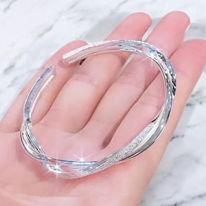 May include: A silver cuff bracelet with a twisted design. The bracelet features a textured, sparkling surface. The open design allows for easy wear. The jewellery is displayed on a hand.