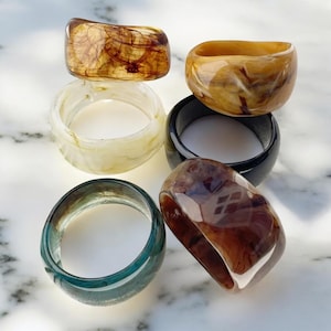 May include: A selection of resin rings in diverse colours and patterns. The rings feature shades of brown, amber, cream, black, and teal. Each ring has a unique marbled or swirled design, highlighting the translucent quality of the resin.