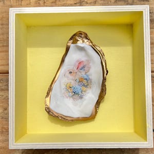 May include: A framed oyster shell art piece. The shell, with a gold-painted edge, features a watercolor painting of a rabbit with a floral arrangement. The artwork is set against a yellow background within a white wooden frame.