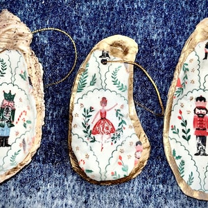 Pode incluir: Três enfeites de Natal de concha de ostra com acabamento dourado e laços para pendurar. Cada concha é decorada com um design diferente do Quebra-Nozes, incluindo uma bailarina e soldados de brinquedo, sobre um fundo branco com detalhes verdes.