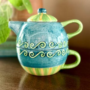 May include: A decorative ceramic teapot and teacup set. The teapot is teal with green and yellow accents, including a striped band and a swirl design. The lid is green and yellow striped. The teacup is stacked beneath the teapot.