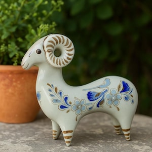 Może przedstawiać: Ceramiczna figurka barana w kremowym kolorze, ozdobiona niebieskimi i brązowymi wzorami kwiatowymi. Baran ma duże, spiralne rogi i stoi na kamiennej powierzchni. W tle doniczka z terakoty i zieleń.