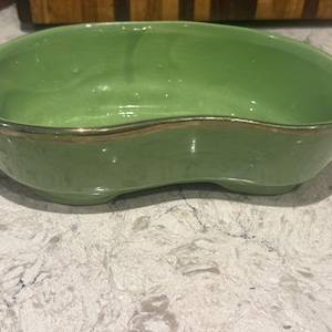 Könnte beinhalten: Eine grüne, nierenförmige Keramikschale mit goldenem Rand. Die Schale hat eine glänzende Oberfläche und steht auf einer hellen Oberfläche. Die Schale ist leer und scheint Vintage zu sein.