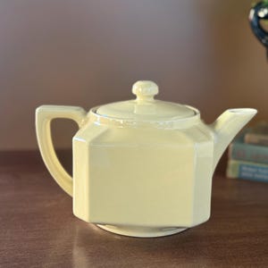 Puede incluir: Una tetera de cerámica de color amarillo pálido con tapa, asa y boquilla. La tetera tiene una forma geométrica octogonal. El asa tiene forma cuadrada. La tetera está sobre una superficie marrón.