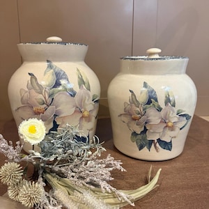 Peut inclure: Deux pots en céramique blanc cassé avec couvercles, ornés d'un motif floral de magnolia dans les tons de violet, bleu et vert. Les pots ont un bord rustique et texturé et sont présentés avec un arrangement floral décoratif.