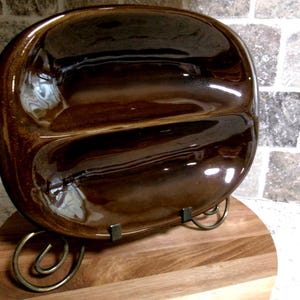 Könnte beinhalten: Eine braune, geteilte Servierschale mit glänzendem Finish. Die ovale Schale steht auf einem Holzsockel mit einem dekorativen Metallständer. Die Schale ist für zwei separate Lebensmittel konzipiert.