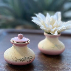 Könnte beinhalten: Zwei kleine Keramikvasen in Rosa und Creme. Eine hat einen Deckel und Blumendetails, die andere hält eine weiße Blume. Die Vasen stehen auf einer dunklen Holzoberfläche. Dekorative Vasen.