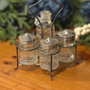 May include: A vintage glass condiment set with a metal stand. The set includes two salt shakers, a pepper shaker, and a small bottle. The glass pieces have etched floral designs and are topped with silver-colored lids.