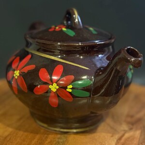 Könnte beinhalten: Eine braune Keramik-Teekanne mit Deckel, Ausguss und Henkel. Die Teekanne ist mit roten und gelben Blumenmustern und grünen Blättern verziert. Auf der Seite der Teekanne ist eine goldene Akzentlinie aufgemalt.