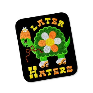 May include: Embroidered patch featuring a green turtle wearing a roller skate and a flower hat. The turtle has a flower-shaped shell and is on roller skates. The words "LATER HATERS" are in yellow and orange.