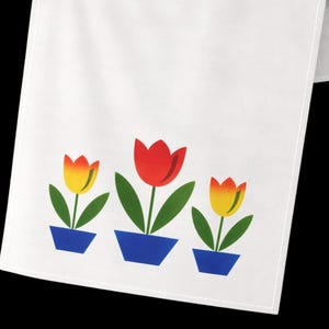 May include: A white fabric table runner featuring a graphic print of three potted tulips. The center tulip is red, while the two flanking tulips are yellow with orange tips. Each flower is set in a solid blue trapezoidal pot with green leaves.