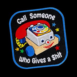 May include: A square embroidered patch with rounded corners and a blue border. It features a vintage toy telephone with a red receiver, a white base, and blue wheels. The text "Call Someone Who Gives a Shit" is embroidered in white.