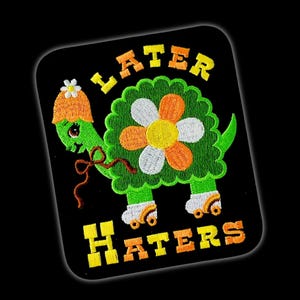 May include: A square embroidered patch on a black background featuring a green turtle wearing an orange hat with a white flower. The turtle has a large flower on its shell and is wearing roller skates. The text LATER HATERS is embroidered in yellow and orange.