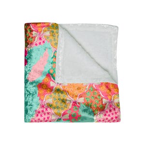 May include: A colorful blanket with a patchwork design. The blanket features a variety of patterns, including bows, stars, and floral motifs in shades of pink, orange, green, and teal. The reverse side is a soft, white, plush material.