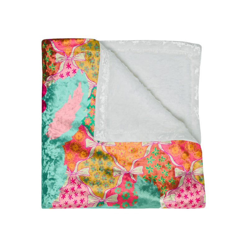 May include: A colorful blanket with a patchwork design. The blanket features a variety of patterns, including bows, stars, and floral motifs in shades of pink, orange, green, and teal. The reverse side is a soft, white, plush material.