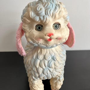 May include: A vintage lamb toy with a cream-colored body and blue accents on its fur. The lamb has pink ears, a red nose, and large, expressive eyes. The toy is made of a soft material and stands upright.