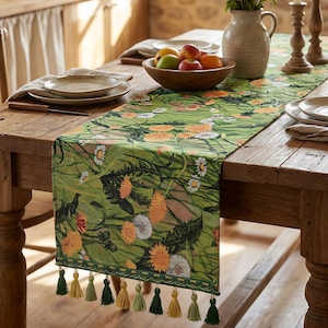 Pode incluir: Caminho de mesa verde com um design floral de flores amarelas e brancas e folhagem verde. O caminho de mesa é decorado com borlas em tons de verde e amarelo. Está sobre uma mesa de madeira com uma tigela de fruta e pratos.