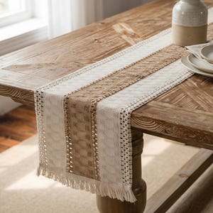 Può includere: Runner da tavolo beige e bianco con bordo a frange. Il runner presenta un design intrecciato con strisce beige e bianche alternate. Il runner è su un tavolo di legno.