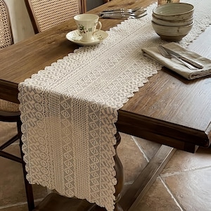 Puede incluir: Un camino de mesa de encaje color crema con un patrón geométrico y floral repetitivo, que presenta bordes festoneados. El camino está colocado sobre una mesa de madera marrón oscuro, acompañado de una taza, cuencos y cubiertos.