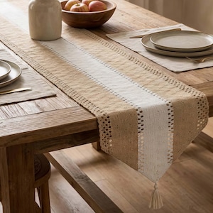 May include: A beige and white striped table runner with a tassel. The runner is made of woven fabric and has a decorative openwork border. The runner is placed on a wooden table, with plates and a bowl of apples visible.