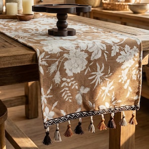 May include: A brown floral table runner featuring white flower and leaf patterns. The runner has a decorative border with tassels in shades of brown and cream. The runner is displayed on a wooden table.