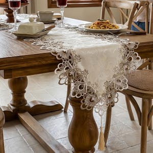 May include: A white table runner featuring an intricate floral lace border in a metallic gold tone. The runner has a smooth central panel with a subtle damask pattern and terminates in a pointed end finished with a long, matching gold tassel.