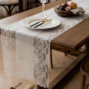 May include: A white lace table runner with a floral pattern and vertical stripes, draped over a wooden table. The runner is set with a white plate, a gold fork, a black knife, a clear glass, and a wooden bowl containing fresh figs and an apple.