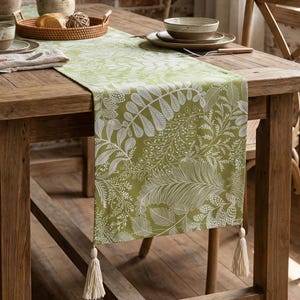 Puede incluir: Camino de mesa verde y blanco con estampado floral y borlas. El corredor presenta un diseño botánico detallado y está sobre una mesa de madera. Las borlas cuelgan de los extremos.