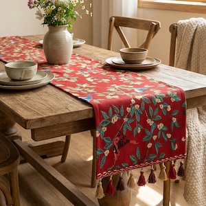 May include: A red table runner with a floral and bird design, featuring green leaves and white flowers. The runner has a tassel fringe and is displayed on a wooden table with tableware and a vase of flowers.