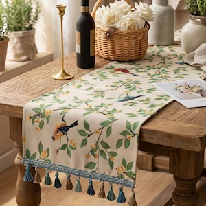 Könnte beinhalten: Beiger Tischläufer mit Vogel- und Blumendesign. Der Läufer hat grüne Blätter, gelbe Blüten und bunte Vögel. Er hat eine blaue Bordüre mit Quasten. Auf einem Holztisch mit Weinflasche, Blumenkorb und Kerzenhalter.