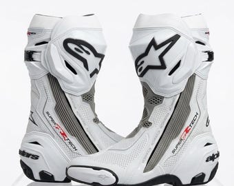 Puede incluir: Un par de botas de moto Alpinestars Supertech blancas con detalles en negro, paneles de cuero perforado y un prominente logotipo de estrella negra en la espinillera. Las botas incluyen secciones flexibles grises y el texto Supertech impreso.