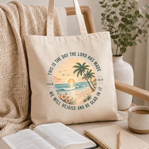 May include: A cream-colored canvas tote bag featuring a circular graphic of a beach scene with palm trees, a sunset, and ocean waves. The text reads: This is the day the Lord has made, Psalm 118:24, we will rejoice and be glad in it.