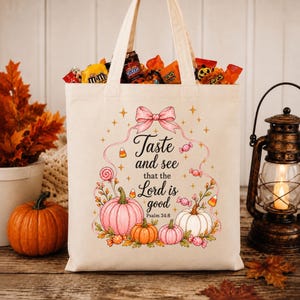 Peut inclure: Un sac fourre-tout en toile beige rempli de bonbons assortis, orné d'un motif imprimé de trois citrouilles, de bonbons colorés et d'un nœud rose. Le texte indique : Taste and see that the Lord is good Psalm 34:8, entouré de petites étoiles dorées.