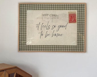May include: A framed wall art piece featuring a vintage postcard design with a green and white plaid border. The center displays the text 'it feels so good to be home' in black script, alongside a postmark and a red postage stamp.