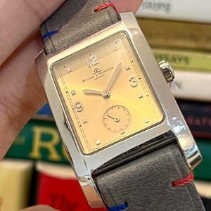Op de afbeelding: Een rechthoekig, zilverkleurig polshorloge met een goudkleurige wijzerplaat en een kleine secondewijzer. Het horloge is voorzien van het merk "Baume & Mercier Geneve". Het heeft een grijze leren band met rode en blauwe stiksels.