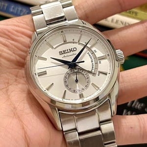 Może przedstawiać: Srebrny zegarek Seiko Automatic z jasną tarczą i bransoletą ze stali nierdzewnej. Zegarek posiada wskaźnik rezerwy chodu i wyświetlacz daty. Tarcza zegarka ma czarne wskazówki i znaczniki. Wyprodukowano w Japonii.