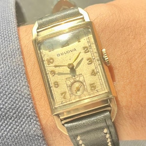 Puede incluir: Un reloj de pulsera Bulova vintage con una caja rectangular dorada y una correa de cuero negro. La esfera del reloj es dorada con números y agujas negras. La esfera del reloj también tiene el texto "Bulova" y "Busch & Sons".