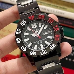 May include: Black Seiko 5 Sports automatic watch with a black dial, white and red accents, and a black metal strap. The watch face displays the day and date, with the text "WATER 100M RESIST AUTOMATIC 23 JEWELS".