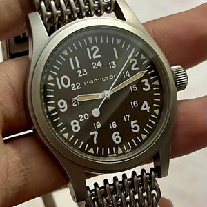 May include: A Hamilton wristwatch with a dark face and white numerals. The watch has a stainless steel mesh strap. The watch face displays the brand name and the word "SWISS MADE".