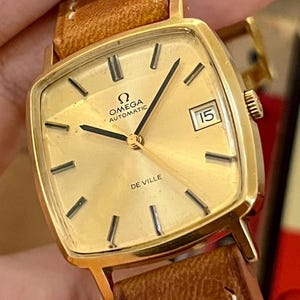 Puede incluir: Un reloj de pulsera automático Omega De Ville dorado con esfera cuadrada y correa de cuero marrón. La esfera del reloj muestra el logotipo de Omega, la palabra "AUTOMATIC" y la fecha. Las manecillas del reloj son negras.