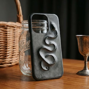 May include: A black phone case with a raised, serpentine design. The case is made of a textured material and features a cutout for the camera. The case is displayed on a wooden surface with a mason jar and a wicker basket in the background.