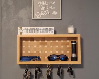 May include: A wooden wall-mounted key holder with a shelf, featuring a woven pattern. The shelf holds sunglasses, a flashlight, and a dog collar. Below, multiple keys hang from hooks. A sign above reads "You, me & the SEA."