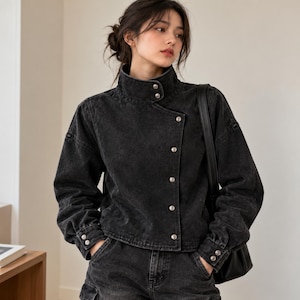 May include: A charcoal black denim jacket featuring a high stand-up collar, long sleeves with buttoned cuffs, and an asymmetrical front closure secured by a vertical row of silver-toned metal buttons. The jacket has a structured, cropped fit.