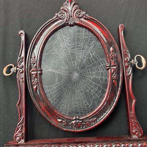 May include: A dark red, oval-shaped tabletop mirror frame featuring ornate, carved floral details and a central glass pane displaying a white spiderweb pattern. The frame is supported by two vertical side pillars and a rectangular base with decorative trim.