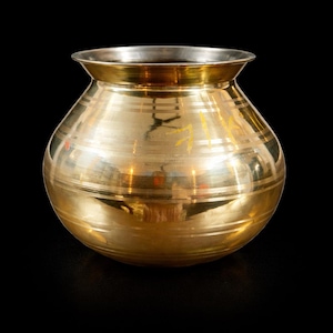 May include: A polished brass pot with a rounded body and a wide, flared rim. The exterior surface features horizontal engraved lines circling the middle and upper sections, with faint yellow markings visible on the side of the metallic vessel.