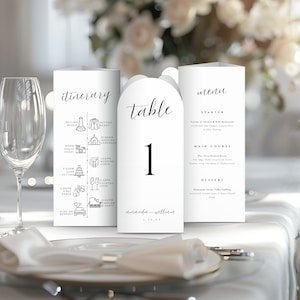 Puede incluir: Elegante juego de papelería de boda blanca. Incluye un itinerario, una tarjeta con el número de mesa 1 y un menú. Las tarjetas presentan caligrafía negra y están colocadas sobre un mantel blanco, con flores en el fondo.