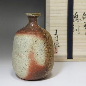 May include: A handcrafted ceramic sake bottle. The bottle has a speckled, off-white body and a reddish-brown top. It has a rounded shape and a narrow neck. The background includes a wooden box with Japanese calligraphy.