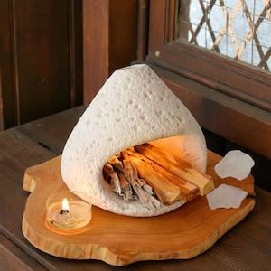 Könnte beinhalten: Ein weißer, strukturierter Keramik-Räuchergefäß in Tropfenform mit Holzsockel. Im Inneren des Brenners befinden sich Holzstücke. Eine kleine Kerze in einem Glashalter steht auf dem Holzsockel. Zwei weiße Steine liegen ebenfalls auf dem Sockel.