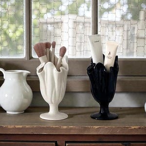 Puede incluir: Una colección de recipientes de cerámica decorativos y brochas de maquillaje sobre una superficie de madera. Los recipientes incluyen una jarra blanca, un jarrón blanco con brochas y un jarrón negro con tubos de crema. El fondo presenta una ventana con malla de alambre.